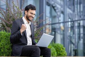 A businessman in a suit celebrates a successful project result while working on his laptop outdoors near an office building. photo