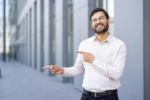 A smiling man with glasses stands outside, pointing towards something. He is wearing a white shirt and looking directly at the viewer. The background has a modern building. photo