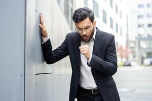 A concerned businessman in a suit leans against a wall, looking distressed or unwell in an outdoor setting, with a hint of anxiety. photo