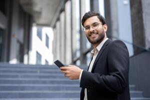 A smiling businessman, dressed in a suit, holding a phone, looks at the camera while standing outside a modern building. photo