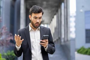 A businessman in a suit appears to be shocked while looking at his phone outdoors. He holds the phone in his hand and reacts with surprise, in front of a building. photo