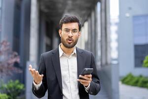 A businessman, dressed in a suit, looks surprised and animated while holding a phone, gesturing with his hands in an outdoor setting. photo