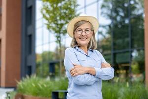 A smiling mature woman stands confidently with crossed arms and hat, in front of a modern building and lush greenery outdoors. photo