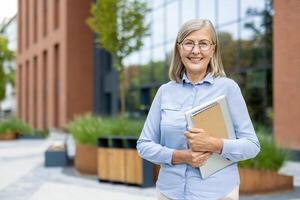 A smiling, mature professional woman stands confidently outside an office building holding a laptop and notebook, showcasing her preparedness. photo