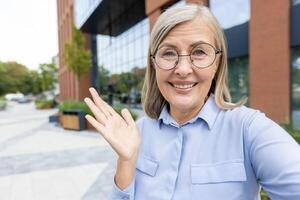 An older woman, wearing round glasses, smiles and gestures at the camera outdoors near a modern building with glass windows. photo