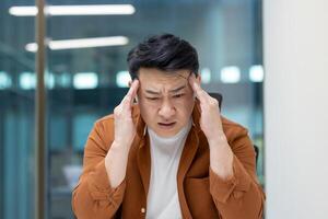 Headache in office worker at workplace. Man suffering from migraine while working inside office. photo