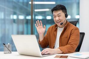 A cheerful asian businessman is waving during a call, looking focused. He is using a headset and laptop in an office setting. photo