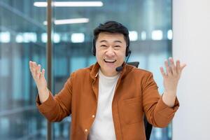Asian man wearing a headset, smiling and gesturing in an office setting. He appears to be in a customer service or call center role, happy. photo