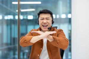 Webcam view, Asian man with headset quarreling angry shouting at camera. Dissatisfied support worker can't understand client. Man at workplace inside office. photo