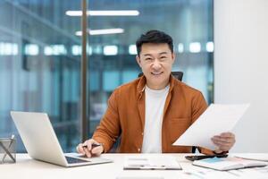Asian businessman smiling while reviewing paperwork at his desk with a laptop in a modern office setting, creating a professional and productive atmosphere photo
