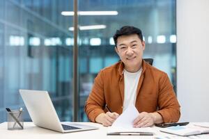 An asian businessman smiles at the camera while working on documents at his desk. He is in a modern office setting, well-lit. photo