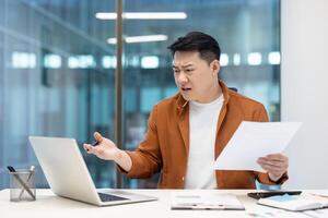 Disappointed and upset financier at workplace inside office. Asian man dissatisfied with report working with documents and papers, using laptop. photo