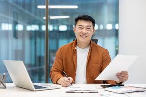 Portrait of successful Asian businessman financier at workplace inside office. Man behind paperwork looking at camera with satisfaction, working with laptop, preparing reports. photo