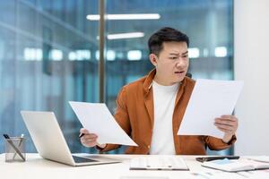 Upset and worried financier looking and reading negative financial reports. Asian nervous man working with documents and papers inside office at workplace. Disappointed businessman. photo
