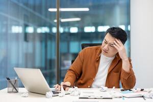 An asian businessman looks stressed while working at his desk, with crumpled paper surrounding him, symbolizing writer's block. photo