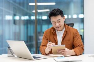 An asian businessman sits at his desk, making notes in a notebook while working on a laptop in a modern office setting. photo