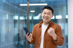An enthusiastic asian man celebrates while holding his smartphone, showing a joyful expression. The man is inside an office setting, holding his phone with a joyful expression. photo