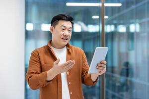 Successful Asian businessman talking to colleagues and partners using call and tablet computer. Man standing near window inside company office. photo