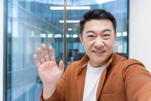 An asian man with a warm smile waves at the camera, seemingly in an office setting with glass windows in the background. photo