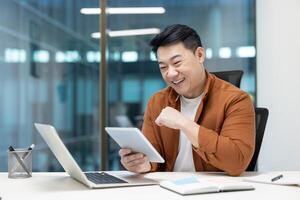 A cheerful programmer enjoys testing an application on a tablet, seated at a modern office desk with a laptop photo