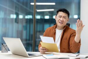 An asian businessman appears perplexed, holding documents in the office, likely facing a work-related problem. photo