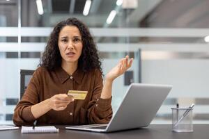 A frustrated woman holds a credit card, appearing perplexed while using a laptop in a modern office setting. She is experiencing an online issue. photo