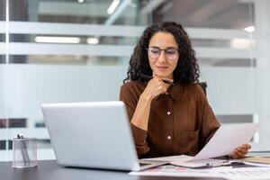 A woman with glasses, working in an office setting, thoughtfully examines documents while using a laptop. photo