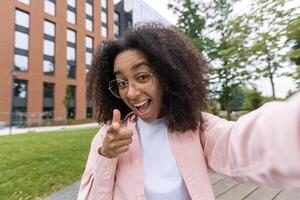 Smiling woman taking selfie outdoors with cheerful expression pointing at camera. Urban setting with trees and building adds vibrant atmosphere. Captures joy, confidence, and connection photo