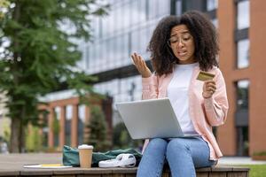 Confused young woman using laptop computer and credit card in outdoor setting. Displaying frustration during online shopping transaction on campus bench. Trees and modern building in background. photo