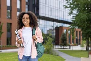 Smiling african american student with glasses and backpack standing on college campus. Holding books and looking at camera. Modern building and greenery in background. photo