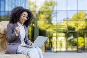 Hispanic businesswoman expressing excitement and success while working on laptop. Seated on bench in front of large glass building. Professional attire, sense of accomplishment and joy. photo