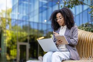 African American businesswoman participates in an online course outside. She engages with instructor via laptop, takes notes in notebook uses for call, enhancing professional skills photo