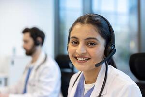 A friendly healthcare professional smiles at the camera while working, wearing a headset in a bright office environment. Another doctor is in the background. photo