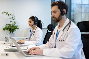 Two healthcare professionals wearing headsets and lab coats, working on laptops in a modern office setting. They are smiling while using technology. photo