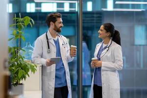 Two doctors, a man and a woman, are chatting amicably during a lunch break, smiling happily, holding cups with a hot drink in their hands, working inside the office of a modern clinic. photo