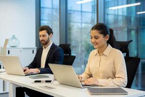 Two colleagues working on laptops in a modern office setting, teamwork concept. The scene shows collaboration and productivity. photo