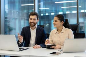 A man and a woman are working together in an office setting, collaborating on a project using their laptops and taking notes. photo