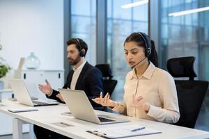 Two office workers wearing headsets are on a call in an open office space, demonstrating remote communication and customer service. photo