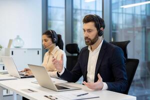 Two business professionals wearing headsets in an office setting, working on laptops with serious expressions. They appear to be customer service agents or call center representatives. photo