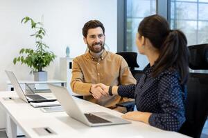 A man and a woman shake hands in a modern office setting with laptops on the desk, symbolizing a business agreement or partnership. photo