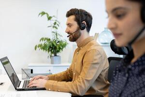 A man with a headset is working on his laptop in a bright office with another colleague working blurred in the foreground. photo