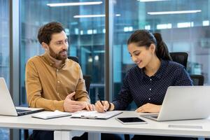 A man and a woman are collaborating in an office setting, with the woman signing documents. They appear to be reviewing a report. photo
