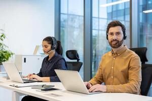 Portrait of customer service customer support worker, man with headset smiling and looking at camera, team working inside office building with laptop. photo