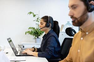 A call center employee with a headset is working on a laptop in a bright office environment. Another person is partially visible. photo