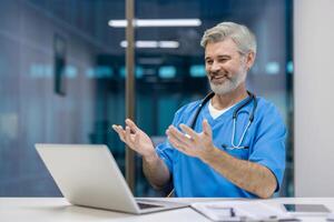 A smiling doctor with a stethoscope is engaged in a call, gesturing with his hands while sitting at a desk with a laptop in an office setting. photo