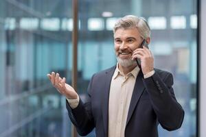 A senior executive in a suit smiles as he talks on his mobile phone in a modern office environment. He gestures with his hand. photo