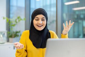A cheerful woman in a hijab gestures enthusiastically while sitting in front of a laptop, likely in an office setting, with an excited expression. photo