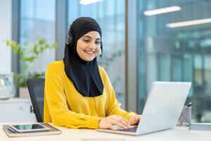A smiling woman wearing a hijab and headset works at a desk, using a laptop in an office setting. The background features a window and office decor. photo