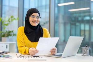 A smiling woman in a hijab works at a desk with a laptop and documents, appearing focused and happy in a modern office environment. photo