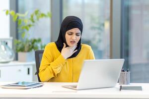 A woman wearing a hijab appears to be suffering from a sore throat while working on a laptop at her desk. She has a concerned expression on her face. photo